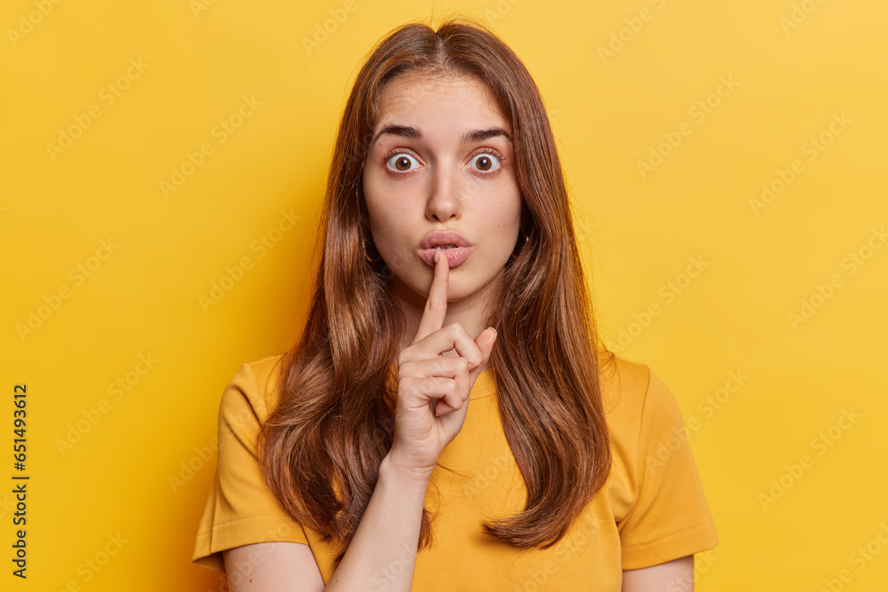 Studio close up of young beautiful European lady with long loose brunette hair in yellow t shirt standing in centre isolated making gesture to keep silence and be quiet and still looking straight - obrazy, fototapety, plakaty 