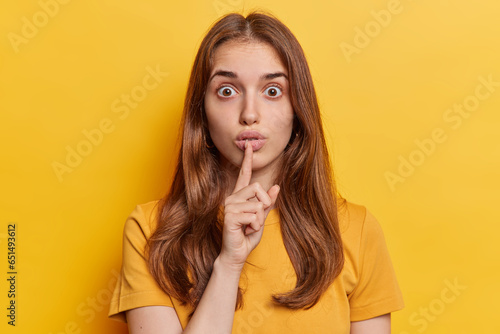 Studio close up of young beautiful European lady with long loose brunette hair in yellow t shirt standing in centre isolated making gesture to keep silence and be quiet and still looking straight