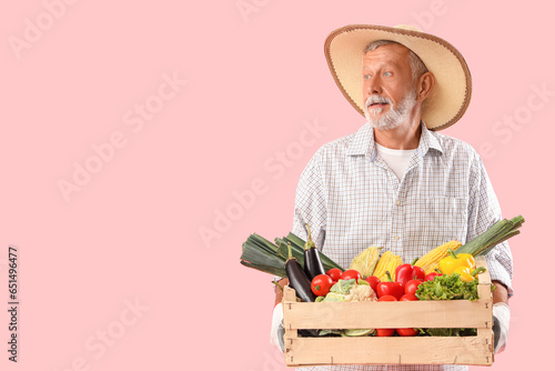 Mature male farmer with wooden box full of different ripe vegetables on pink background
