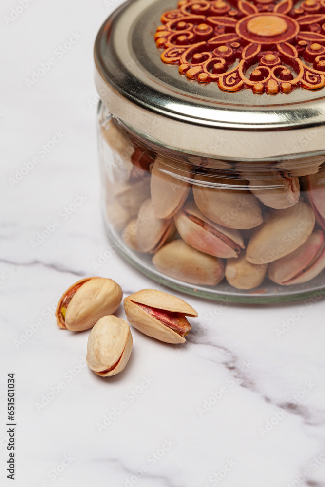 Fresh salted Pistachio nuts into a glass jar with a designer lid, isolated on a marble background. Front view.