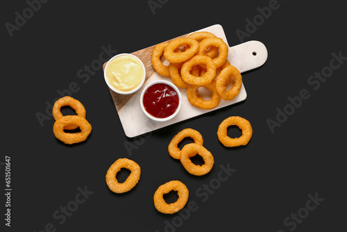 Board with fried breaded onion rings and different sauces on black background