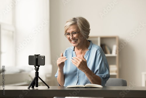 Cheerful senior elder influencer woman working on shooting for blog, recording selfie video at home, speaking at cellphone camera fixed on tripod, smiling, broadcasting online