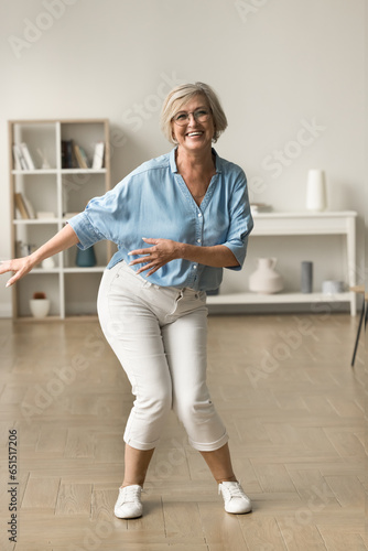 Cheerful carefree active retired woman dancing at home, enjoying motion, leisure, music, relaxation, looking at camera, posing for full length indoor portrait, having fun, laughing photo