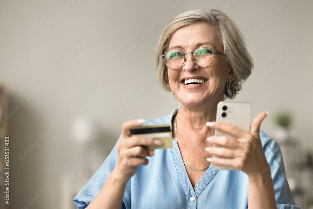 Cheerful confident elderly woman in elegant eyeglasses holding credit card and smartphone, looking away, smiling, laughing, enjoying online shopping on sale, using commerce application