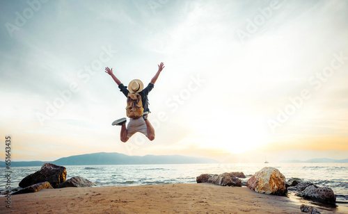 Happy man with backpack standing with arms up at the beach - Delightful tourist enjoying summer vacation by the seaside - Traveling life style and well being concept
