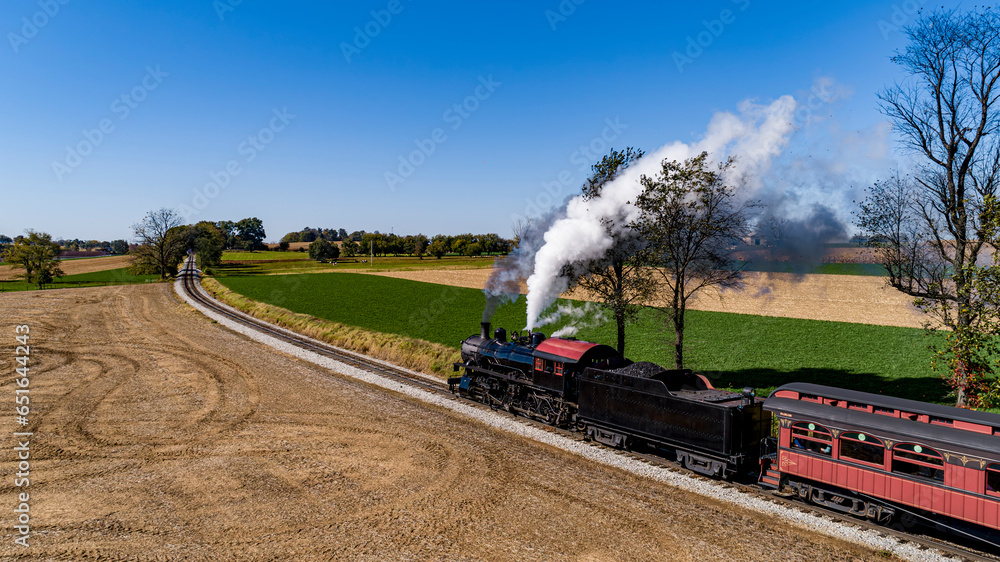 Obraz premium A Drone Side View of an Antique Steam Passenger Train, Rounding a Curve, Blowing Smoke, on a Sunny Fall Day