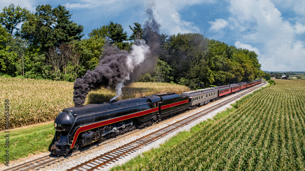 An Aerial Front View of a Restored Antique Steam Passenger Train, Traveling Thru the Countryside, Passing Corn Fields and Blowing Smoke on a Sunny Summer Day - obrazy, fototapety, plakaty 