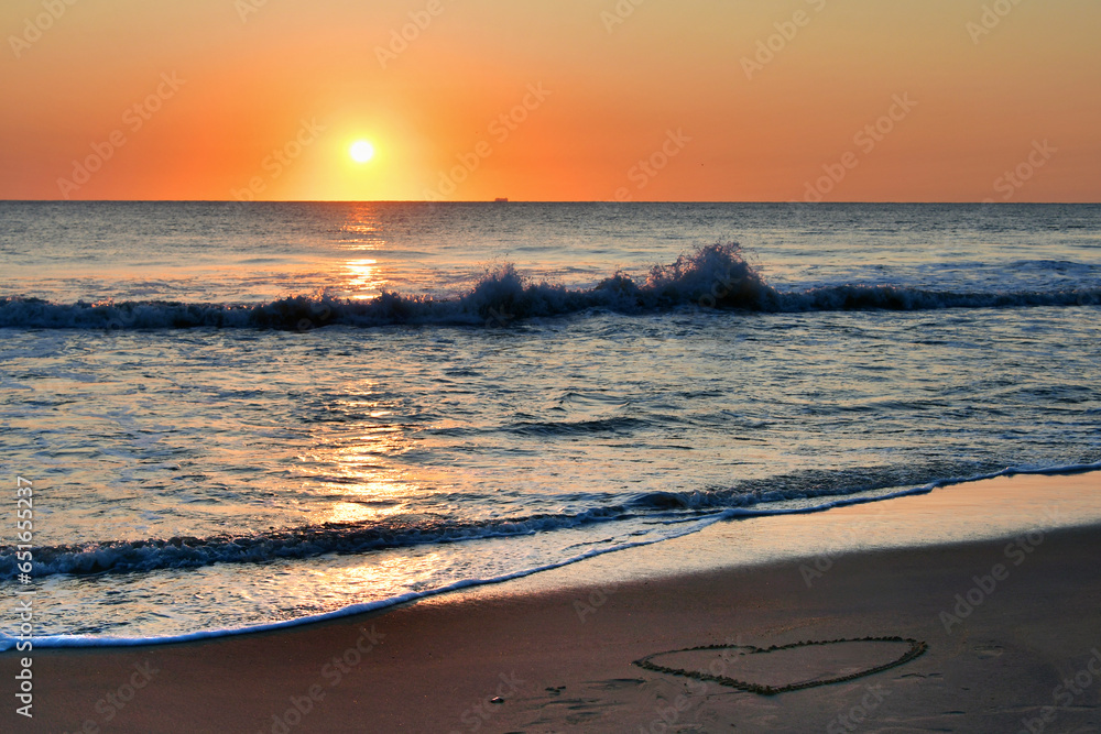 Fototapeta premium Heart drawn in the sand on the beach at sunrise over the ocean