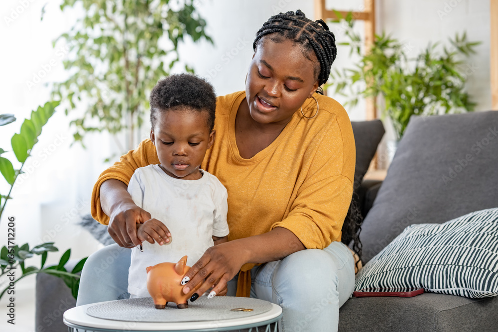 Cute little African-American boy with his mother inserting a coin into a piggy bank, financial and banking concept. Child saving money for the future concept.