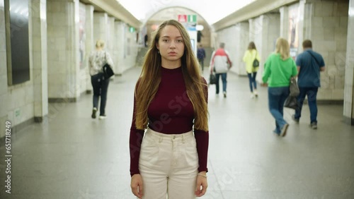 A young beautiful girl with long hair in white jeans and a burgundy sweater stands motionless and looks at the camera, and then closes her eyes in subway while people behind her are walking hurriedly