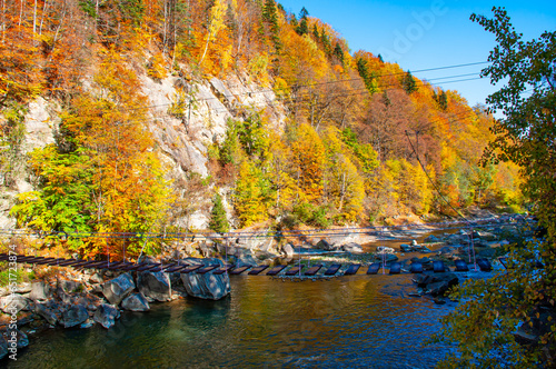Rocky coast with autumn trees in the sun over a mountain river. Suspension bridge over the mountain