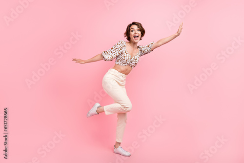 Full length photo of funny optimistic woman wear print top white pants playing hold hands like wings isolated on pink color background