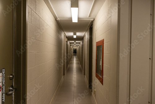 A long, narrow hallway with closed gray doors and a red fire extinguisher - white walls and tiled floor - fluorescent lights overhead. Taken in Toronto, Canada.