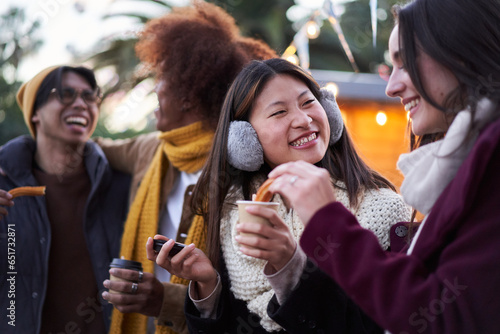 Happy smiling friends eating chocolate with churros together on the street outdoors. Tour group of four multiracial people gathered having fun at festival in winter. Persons holding cup of hot drink. 