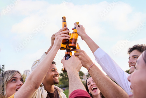 Cheerful young friends toasting together with beer. Happy diverse people clinking bottles outdoors at spring party in the park. Multiracial group having fun and celebrating friendship in community. 