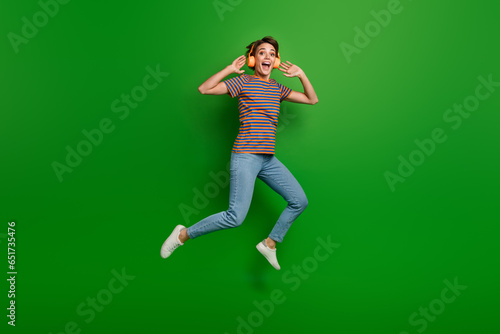 Full body photo of pretty overjoyed astonished lady jumping listen favorite song isolated on green color background