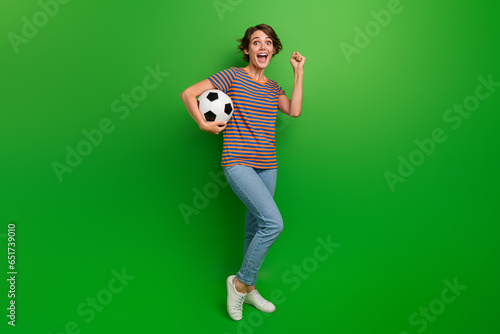 Full size photo of delighted pretty girl hand hold football raise fist success luck isolated on green color background