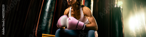 Asian female Muay Thai boxer or kickboxing taking short break sitting with her gloves on at the gym with boxing equipment in background. Strong and muscular body sportswoman. Spur