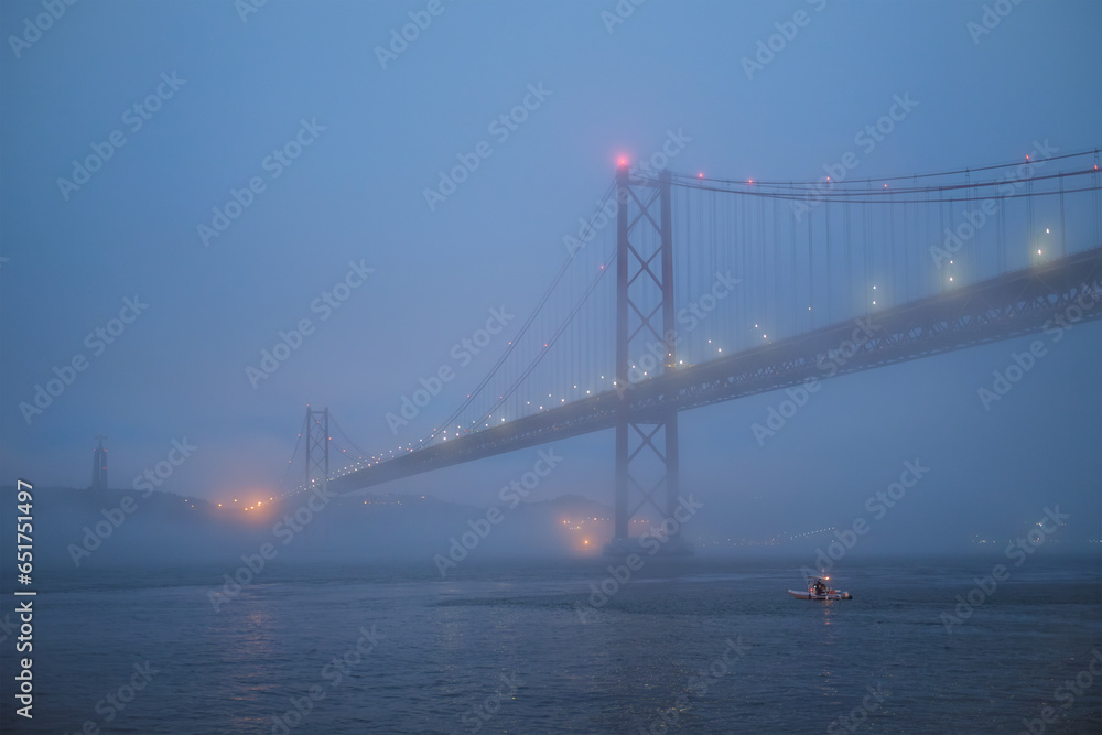 View of 25 de Abril Bridge famous tourist landmark of Lisbon connecting Lisboa and Almada in heavy fog mist wtih yacht boats passing under. Lisbon, Portugal