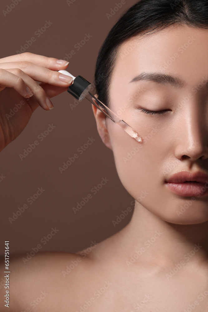 Beautiful young woman applying cosmetic serum onto her face on brown background, closeup Beautiful young woman applying cosmetic serum onto her face on brown background, closeup