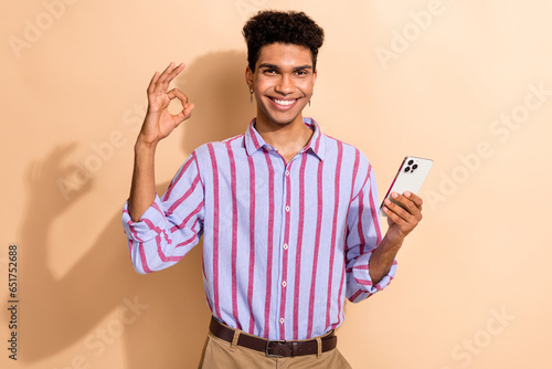 Photo portrait of nice young guy show okey symbol hold device dressed stylish striped formalwear garment isolated on beige color background