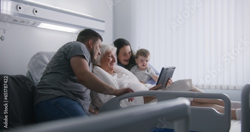 Hospital room. Loving family support elderly grandmother recovering after successful surgery. Relatives talk by video call using digital tablet, spend time together. Modern medical facility, clinic.