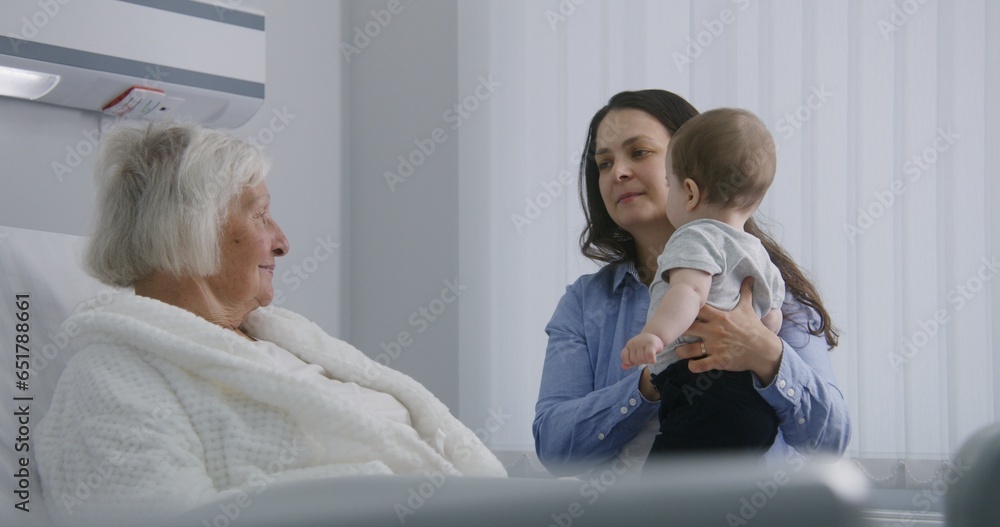 Elderly woman lying and resting in bed in bright hospital ward, plays with grandson. Loving family members support grandmother recovering after successful surgery. Modern medical facility or clinic. Elderly woman lying and resting in bed in bright hospital ward, plays with grandson. Loving family members support grandmother recovering after successful surgery. Modern medical facility or clinic.