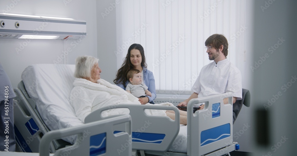 Obraz premium Elderly woman and her family in hospital room. Doctor uses digital tablet, consults female senior patient recovering after successful surgery. Nurse adjusts pillow and bed. Modern medical facility.