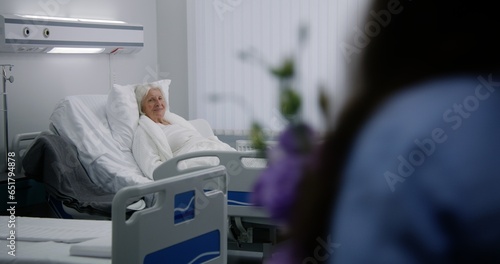 Senior female patient lies and rests in bed in bright hospital ward. Loving family members come to elderly woman recovering after successful surgery, give flowers. Modern medical facility or clinic.