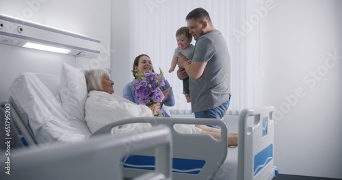 Senior female patient lies and rests in bed in bright hospital ward. Loving family members come to elderly woman recovering after successful surgery  give flowers. Modern medical facility or clinic.