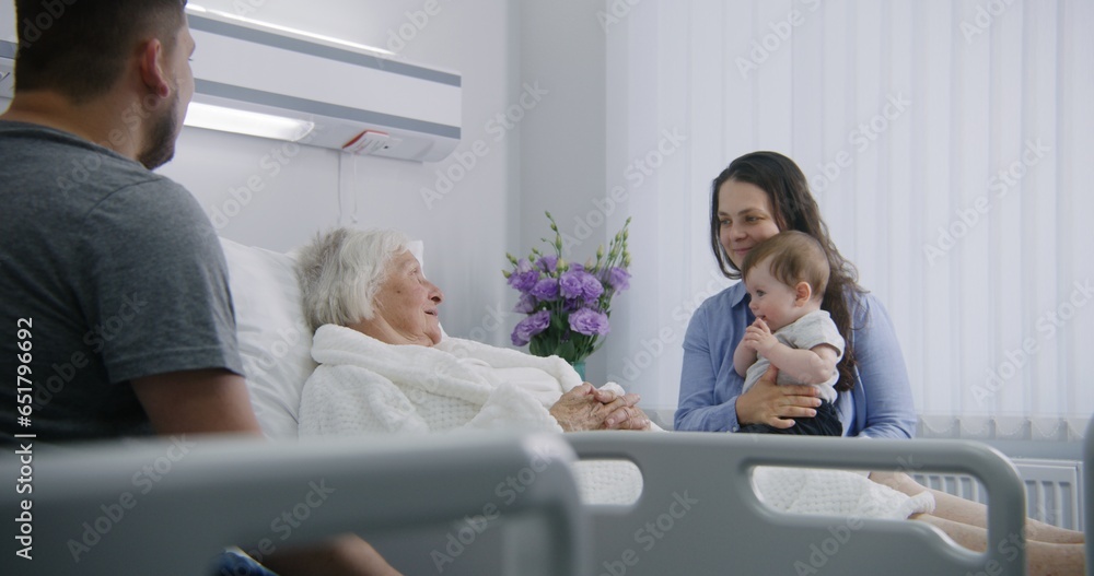 Obraz premium Female elderly patient lies in bed in bright hospital ward, talks to daughter with grandson. Loving family supports senior woman recovering after successful surgery. Modern medical facility or clinic.