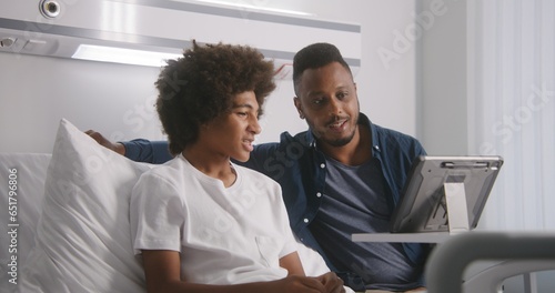 African American teenager rests on bed in hospital ward. Man visits son after surgery. Family remotely talk using tablet computer. Patient fully recovering after successful operation. Modern clinic.