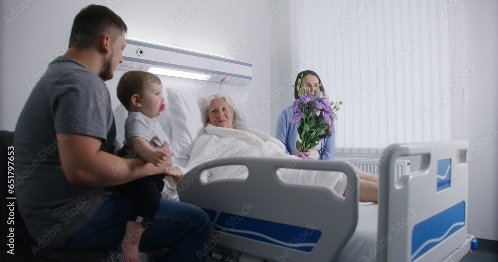 Elderly woman lying and resting in bed in bright hospital ward, plays with grandson. Loving family members support grandmother recovering after successful surgery. Modern medical facility or clinic. Elderly woman lying and resting in bed in bright hospital ward, plays with grandson. Loving family members support grandmother recovering after successful surgery. Modern medical facility or clinic.