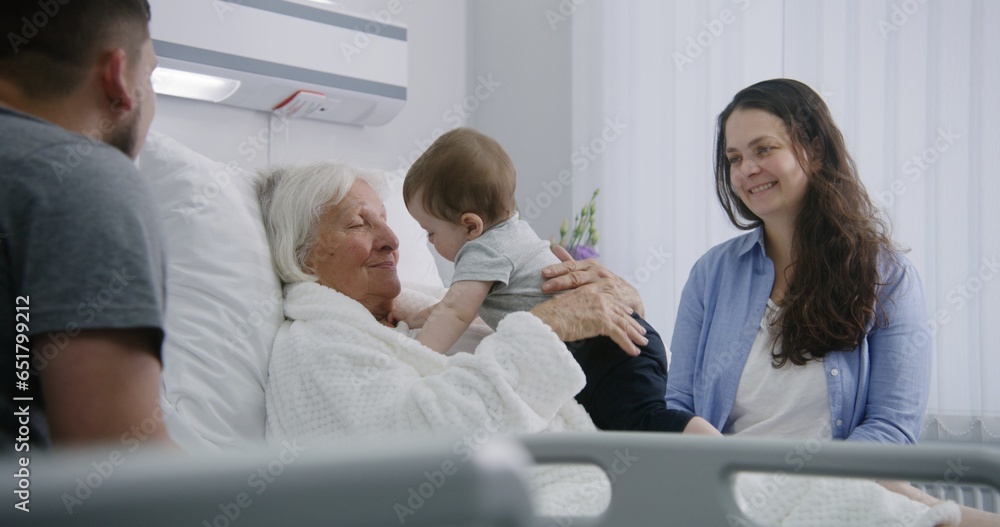 Obraz premium Female elderly patient lies in bed in bright hospital ward, plays with little grandson. Loving family members support senior woman recovering after successful surgery. Modern medical facility.