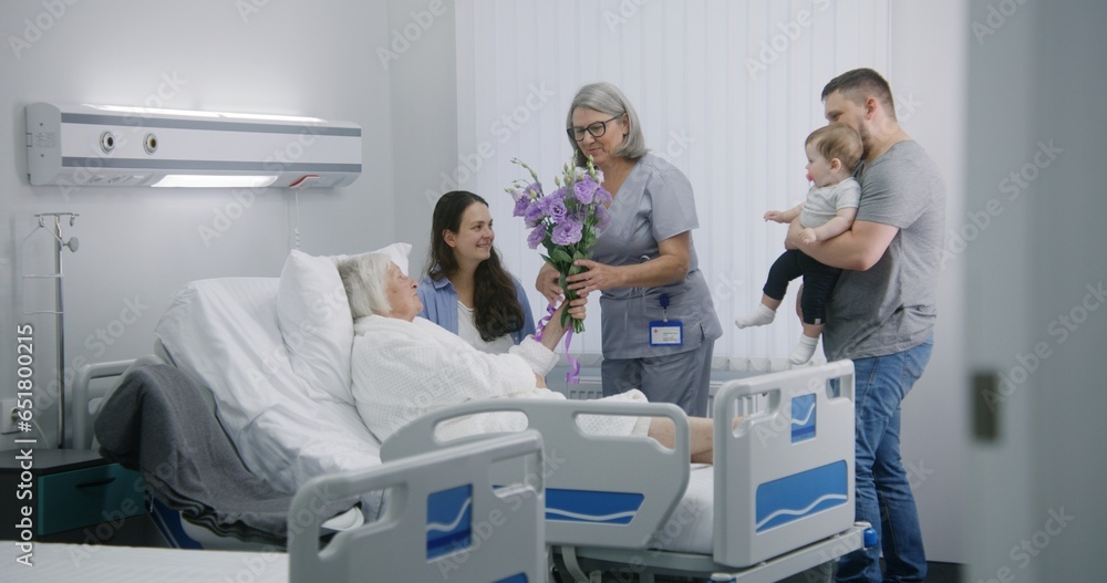 Obraz premium Senior female patient lies and rests in bed in bright hospital ward. Loving family members come to elderly woman recovering after successful surgery, give flowers. Modern medical facility or clinic.