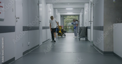 Mature cleaner mops floor in medical center corridor. Multicultural teens stand near window and talk. Caucasian boy with broken leg leaves his friend and goes to hospital ward down clinic hallway.