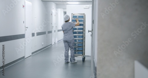 Nurse in uniform pushes mobile shelves with inventory through bright clinic corridor. Female medical worker takes box from rack, enters hospital ward or laboratory. Hallway of modern medical facility.
