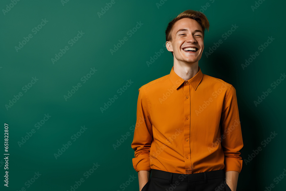 Fototapeta premium Spontaneously laughing young man in orange shirt against green backdrop.
