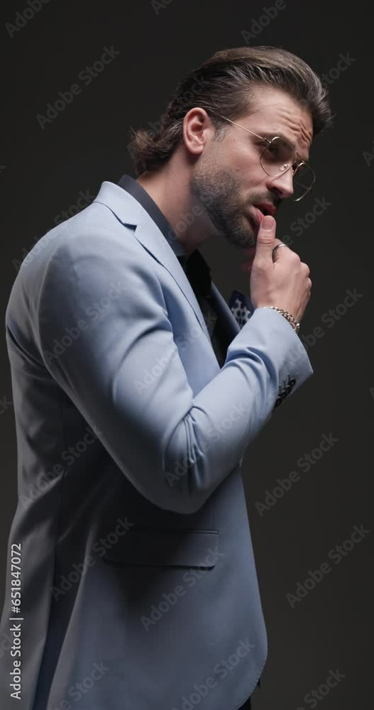 cool fashion guy with glasses looking down and moving, touching lips in a seductive manner, rubbing palms and holding hands in pockets on grey background cool fashion guy with glasses looking down and moving, touching lips in a seductive manner, rubbing palms and holding hands in pockets on grey background