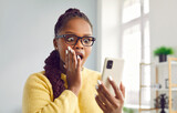 Woman is amazed by content she saw or reads shocking message on mobile phone screen. African American woman opens her mouth in surprise and holds her hand on her cheek while looking at her smartphone.