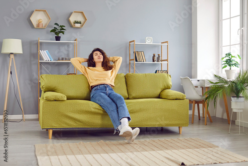 Happy relaxed young woman sitting on comfortable sofa. Beautiful serene girl in casual clothes holding hands behind head resting on couch at home enjoying free time, lazy weekend time