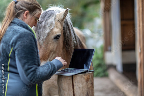 Obraz na płótnie Horse owner as digital online clients: A laptop in front of blurred horse and owner paddock scene, consulting online advice and online shopping as horse owner w salonie