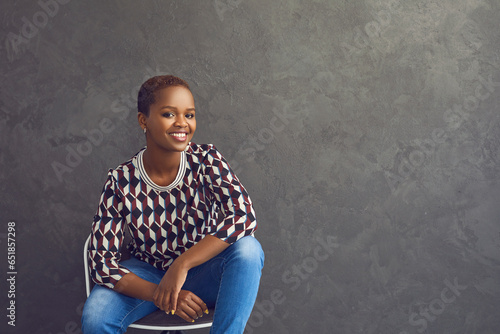 Studio portrait of happy and confident dark skinned casual woman who is smiling while looking at camera. Woman in casual clothes sitting on chair on gray background. Place for text. Advertising banner