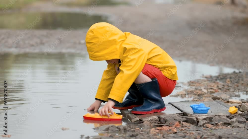 A 3-4 year old boy in a yellow waterproof raincoat and rubber boots launches boats in a puddle in the rain. Children's fun and street games A 3-4 year old boy in a yellow waterproof raincoat and rubber boots launches boats in a puddle in the rain. Children's fun and street games