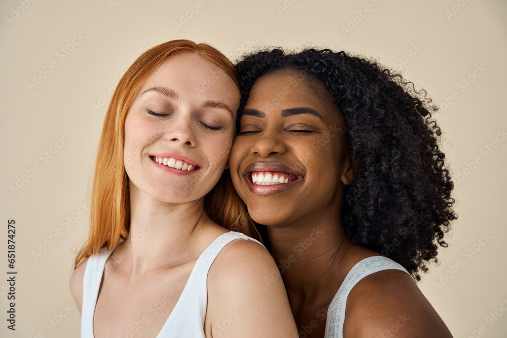 Two smiling diverse cute young girls bonding eyes closed isolated on beige background. Happy African and Caucasian women models pretty faces advertising youth moisturizing skin care natural beauty.