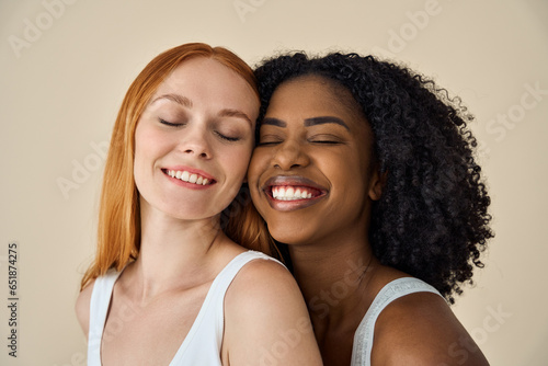 Two smiling diverse cute young girls bonding eyes closed isolated on beige background. Happy African and Caucasian women models pretty faces advertising youth moisturizing skin care natural beauty.