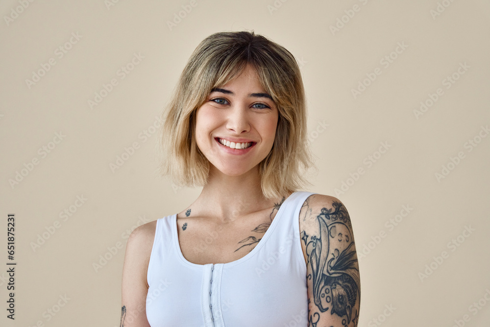 Young happy blond pretty smiling girl beauty female gen z model with short blonde hair beautiful face healthy skin and tattoo looking at camera wearing white top isolated at beige background. Portrait Young happy blond pretty smiling girl beauty female gen z model with short blonde hair beautiful face healthy skin and tattoo looking at camera wearing white top isolated at beige background. Portrait