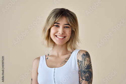 Young happy blond pretty smiling girl beauty female gen z model with short blonde hair beautiful face healthy skin and tattoo looking at camera wearing white top isolated at beige background. Portrait