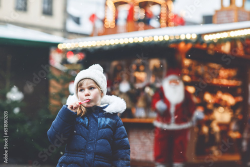 Little cute preschool girl with candy cane from a sweets stand on Christmas market. Happy child on traditional family market in Germany. Preschooler in colorful winter clothes during snowfall