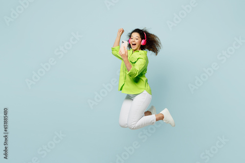 Full body fun young latin woman she wear green shirt casual clothes jump high hold in hand use mobile cell phone isolated on plain pastel light blue cyan background studio portrait. Lifestyle concept.